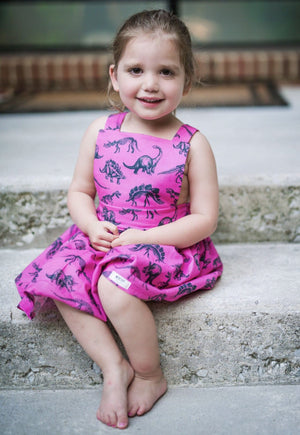 Baby pinafore dress modeled by toddler sitting on step.  Magenta pink dinosaur dress by Worthy Threads, brand for unique baby clothes.