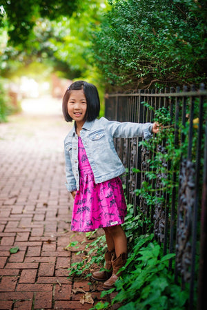 Baby pinafore dress modeled by toddler holding onto a fence: Pink dinosaur dress under a jean jacket.  Girls dinosaur clothes available in matching sister outfits newborn and toddler!